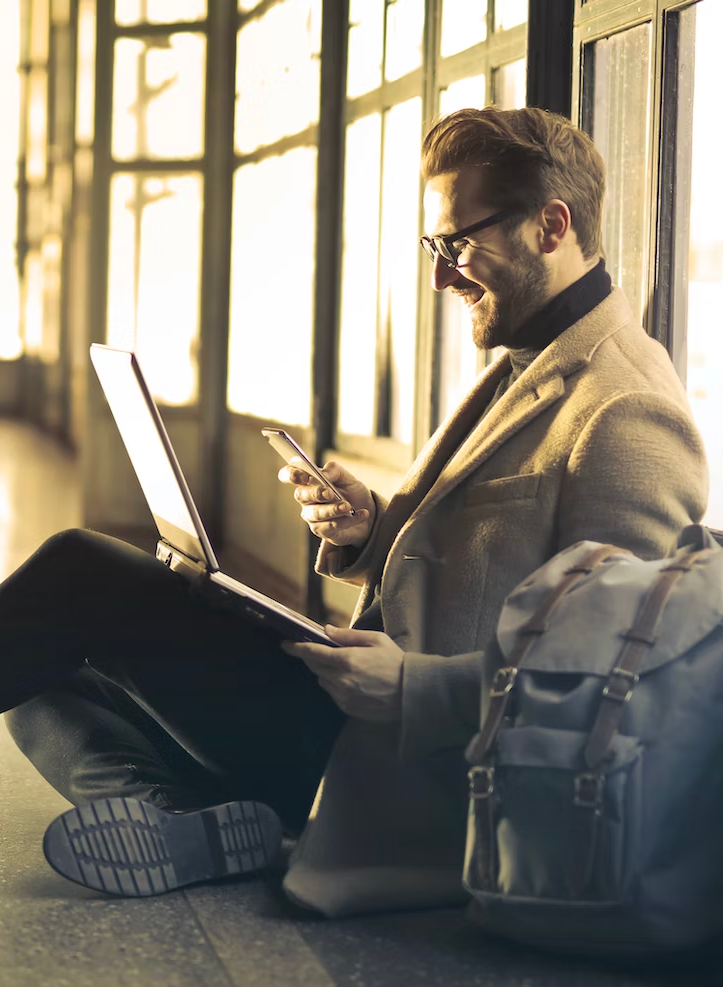 Man wearing a business suit sitting on the corner with a latoptop on his lap while looking at his phone smliling.